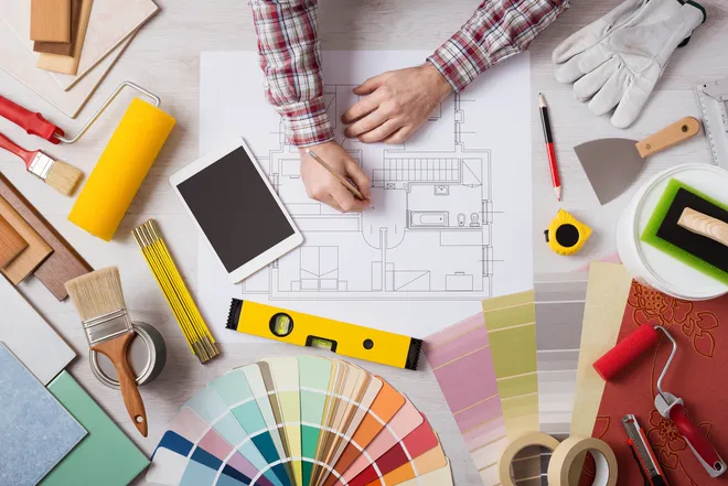 Top-down view of a painter's desk with house plan, paint tools, color swatches, and renovation equipment – home improvement planning and cost estimation.